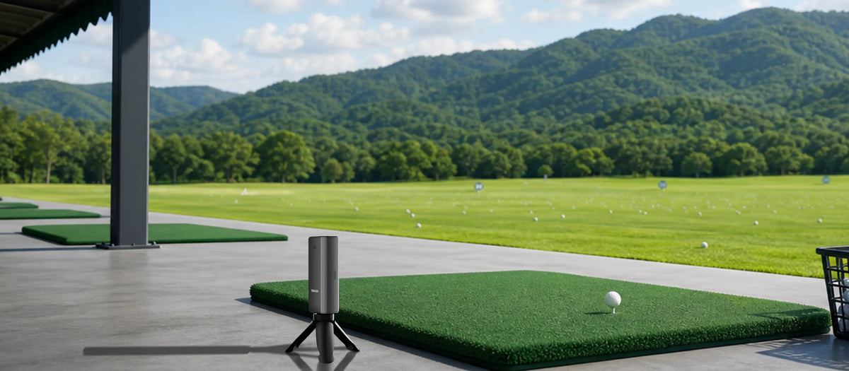 A golf driving range scene with multiple green turf practice mats on a concrete porch, a golf ball on one mat, and a large golf course with trees and mountains.